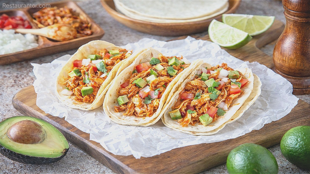 Street-style tacos filled with shredded meat and topped with onion, tomato, cilantro, and avocado, with lime wedges